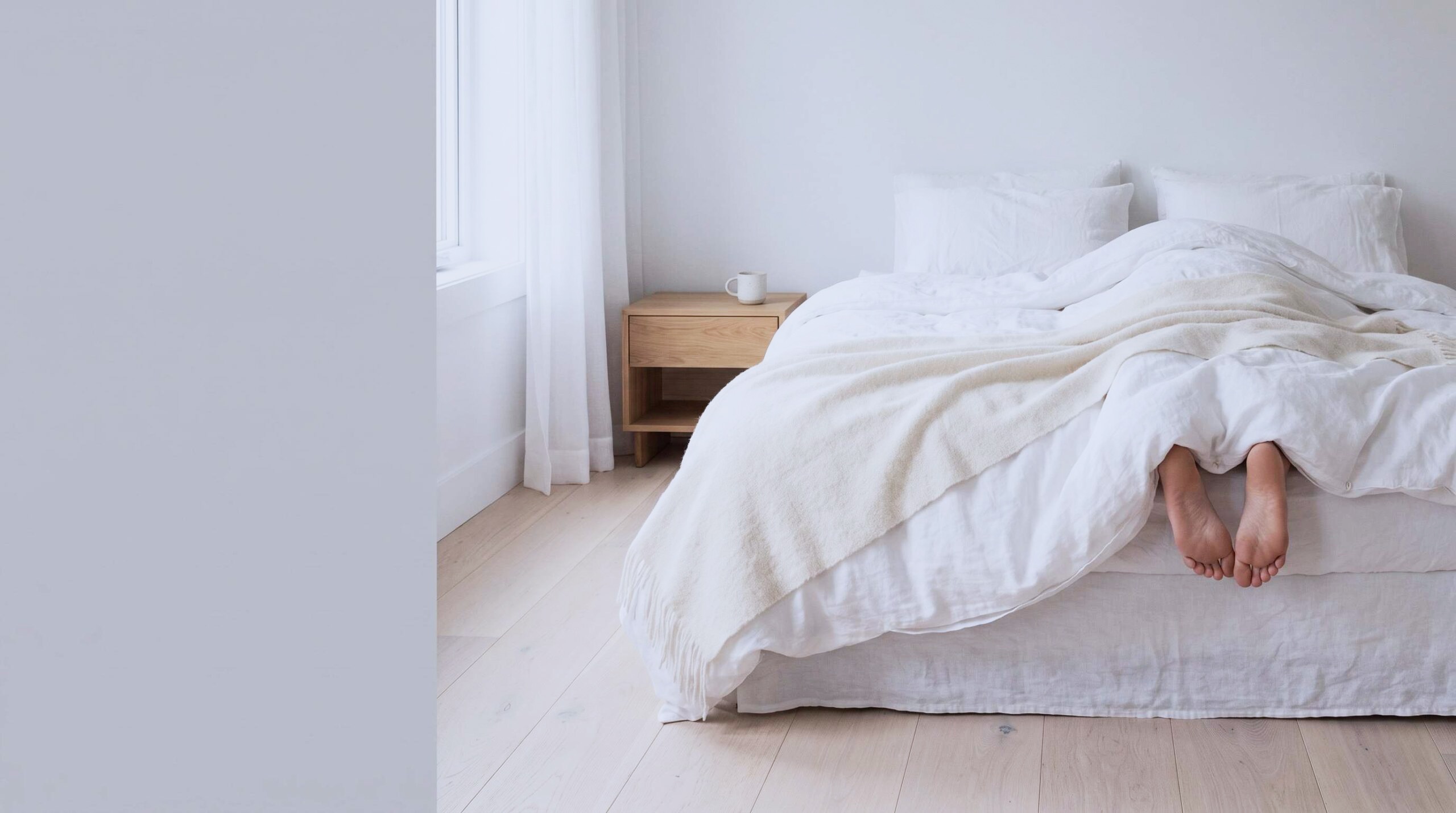 Bare feet peeking out from under a clean white blanket in a bright, airy bedroom